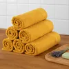 A stack of rolled-up yellow towels on a wooden counter, with a tiled white wall in the background. A small wooden tray with lavender and soap is in the foreground. Produktbild-Vorschau