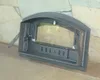 A close-up of a black cast iron pizza oven door with a glass window, resting on a stone hearth in front of a stone wall. Produktbild-Vorschau