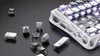 A close-up view of a white mechanical keyboard with transparent keycaps and purple backlighting, surrounded by loose keycaps and switches on a dark, textured surface. Náhled obrázku produktu