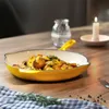 A yellow cast iron pan filled with roasted potatoes and herbs sits on a wooden table, next to a checkered cloth and a lit candle. Produktbild-Vorschau