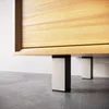 A close-up view of the bottom of a wooden cabinet or furniture piece, supported by two modern, square legs with black tops and light-colored bases, resting on a concrete floor. Produktbild-Vorschau