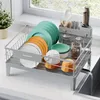 A gray dish rack filled with dishes, bowls, and cups is placed on a kitchen counter next to a sink with water draining. Produktbild-Vorschau