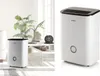A white dehumidifier is shown in a bright, modern living room setting with plants and decorative items. Produktbild-Vorschau
