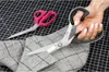 A hand is holding a pair of scissors cutting fabric, with another pair of scissors in the background. Anteprima dell’immagine prodotto