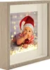 A framed photo of a baby in a Christmas hat holding a bauble, surrounded by fairy lights. Produktbild-Vorschau