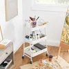 A white three-tier rolling cart filled with art supplies, placed next to a nightstand and an easel with a painting. Náhled obrázku produktu