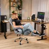 A woman is sitting in a dark gray ergonomic office chair with a footrest extended, in a home office setting. Produktbild-Vorschau