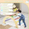 A young child is climbing on a wooden climbing arch with colorful rungs, placed on a soft mat. In the background, there are stuffed toys and a rainbow graphic. Produktbild-Vorschau