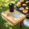 A close-up shot of a wooden cutting board with grilling tools and a black container, placed on a grill with burgers cooking. Podgląd zdjęcia produktu