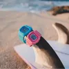 Two G-Shock watches, one blue and one pink, are placed on a surfboard fin on a beach with the ocean in the background. Produktbild-Vorschau