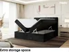 A bedroom scene featuring a black upholstered bed with its storage compartment open, revealing the mattress and topper. The room is elegantly decorated with soft lighting and a large window. Produktbild-Vorschau