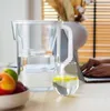 A water filter pitcher and a glass of water are on a table, with a person working on a laptop in the background. Podgląd zdjęcia produktu