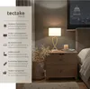 A bedside table with a lamp and a basket of dried flowers, next to a bed, with a city skyline visible through a window. Produktbild-Vorschau