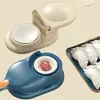 A close-up shot of two dumpling makers, one beige and one blue, with a tray of finished dumplings in the background. Produktbild-Vorschau