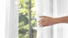 A hand is pulling back a sheer white curtain, revealing a blurred background of green trees and a building. Produktbild-Vorschau