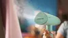A person is holding a mint-colored handheld garment steamer, which is emitting steam. Produktbild-Vorschau