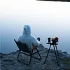 A person in a white hooded sweater sits in a camping chair by a lake, with a small table set up next to them with food and a drink. Produktbild-Vorschau