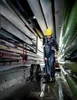 A man wearing a yellow hard hat and work clothes is using a wet/dry vacuum cleaner to clean up a flooded floor in a room with many pipes overhead. Náhled obrázku produktu