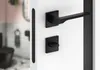 A black door handle set is mounted on a white door, with a blurred view of a bathroom interior in the background. Produktbild-Vorschau