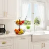 A two-tiered metal fruit basket filled with various fruits sits on a kitchen counter next to a coffee maker and a sink with a window in the background. Produktbild-Vorschau