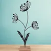 A black metal silhouette of a flower with three seed heads and two leaves stands on a light brown wooden base against a light blue background. Produktbild-Vorschau