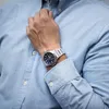 A man wearing a light blue button-down shirt and a silver watch with a blue face. Produktbild-Vorschau