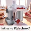 A close-up shot of a meat grinder attachment on a kitchen machine, with raw meat being processed. In the background, a person is preparing more meat. Produktbild-Vorschau