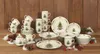 A collection of Christmas-themed tableware, including mugs, plates, and a tiered stand, arranged on a wooden surface with festive decorations like pinecones and berries. Produktbild-Vorschau