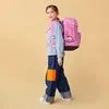A young girl wearing a pink backpack and jeans walks against a plain yellow background. Voir les images du produit