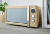 A retro-style cream microwave oven is displayed on a wooden countertop next to a plant and a shelf with books. Náhled obrázku produktu