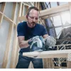 A man is using a circular saw to cut a piece of wood in a construction setting. Produktbild-Vorschau