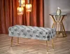 A tufted upholstered bench with gold legs sits in a room with a draped curtain and a side table. Produktbild-Vorschau