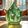 A decorative glass teardrop-shaped lamp with a winter forest scene and deer motif, placed on a wooden table in a living room setting with a sofa and window in the background. Produktbild-Vorschau