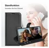 A woman is holding a smartphone in a black wallet case, which is propped up to display a video of a couple. The case is shown in the foreground, with the woman and phone in the background. Produktbild-Vorschau