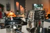 A collection of stainless steel pots and pans are neatly stacked and arranged on a countertop, with a blurred background of a room with decorative lights. Náhled obrázku produktu