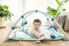 A baby is lying on a play mat with a play gym over them, surrounded by plants and home decor. Produktbild-Vorschau