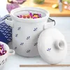 A white ceramic crock with blue floral accents, filled with shredded red cabbage and carrots, sits on a light blue wooden surface next to its lid. A blue and white patterned cloth is visible to the left. Náhled obrázku produktu