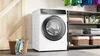 A Bosch washing machine is shown in a laundry room setting, next to a basket of clothes and shelving with folded towels. Produktbild-Vorschau