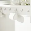 A close-up view of a white wall-mounted shelf with several metal hooks holding white mugs. Produktbild-Vorschau