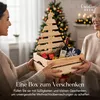 Two people are holding a wooden gift box filled with Christmas-themed items, including a bottle of wine and small wrapped gifts, in front of a decorated Christmas tree. Produktbild-Vorschau