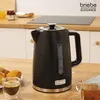 A black electric kettle is shown on a wooden table, with pastries and coffee in the foreground, and kitchen cabinets and a stovetop in the background. Podgląd zdjęcia produktu