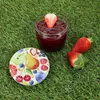 A jar of jam with a strawberry on top, surrounded by fresh strawberries and a decorative lid with fruit illustrations, all placed on a grassy surface. Podgląd zdjęcia produktu