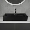 A black ceramic vessel sink with a chrome faucet is shown on a white countertop, with a concrete wall and a circular mirror in the background. Podgląd zdjęcia produktu