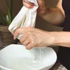 Hands squeezing a white cloth over a white bowl, with liquid dripping into the bowl. Produktbild-Vorschau