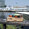 A small pop-pop boat toy sits on a cardboard box on a railing, with a body of water and boats in the background. Produktbild-Vorschau