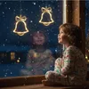 A child in pajamas sits by a window on a snowy night, looking at illuminated Christmas bell decorations hanging outside. Produktbild-Vorschau
