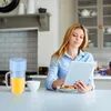 A woman is using a tablet in a kitchen while a portable blender with orange juice is on the counter. Produktbild-Vorschau