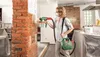A woman is using a paint sprayer on a brick wall in a room that appears to be under renovation. Produktbild-Vorschau
