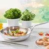 A stainless steel frying pan filled with cooked shrimp and vegetables, placed on a white wooden table with potted herbs and a small bowl of sauce in the background. Anteprima dell’immagine prodotto