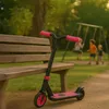A pink and black scooter is parked next to a wooden bench in a park, with children playing on a swing set in the background. Anteprima dell’immagine prodotto